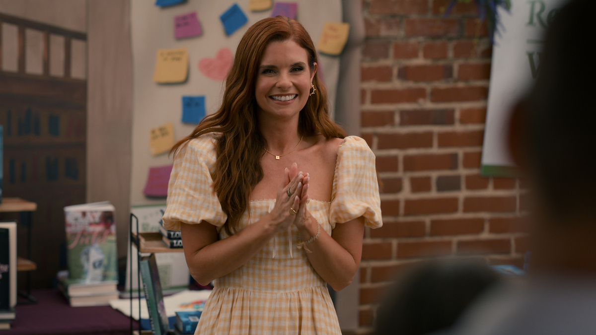 "Woman in a yellow plaid dress smiling and clasping hands at an indoor event, with books, sticky notes, a brick wall, and posters visible in the background."