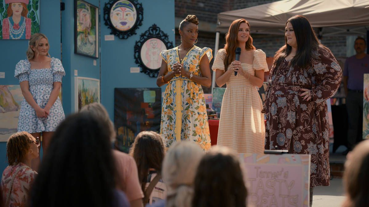 "Four women stand on a stage at an outdoor art fair, three with microphones addressing an audience. Colorful paintings and art are displayed on a blue wall behind them. The crowd is seated in the foreground."