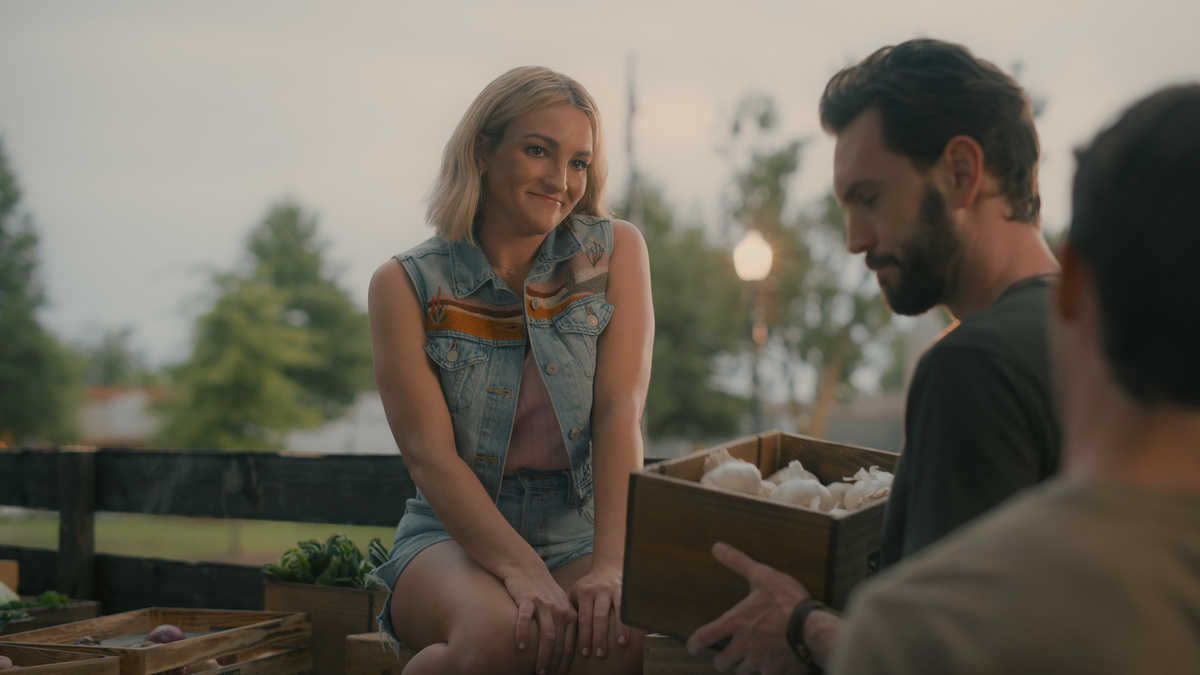 "Woman in a denim vest sits on a wooden surface at an outdoor market while two men handle a box of garlic; fresh produce and trees are visible in the background, suggesting a relaxed, rural community setting."